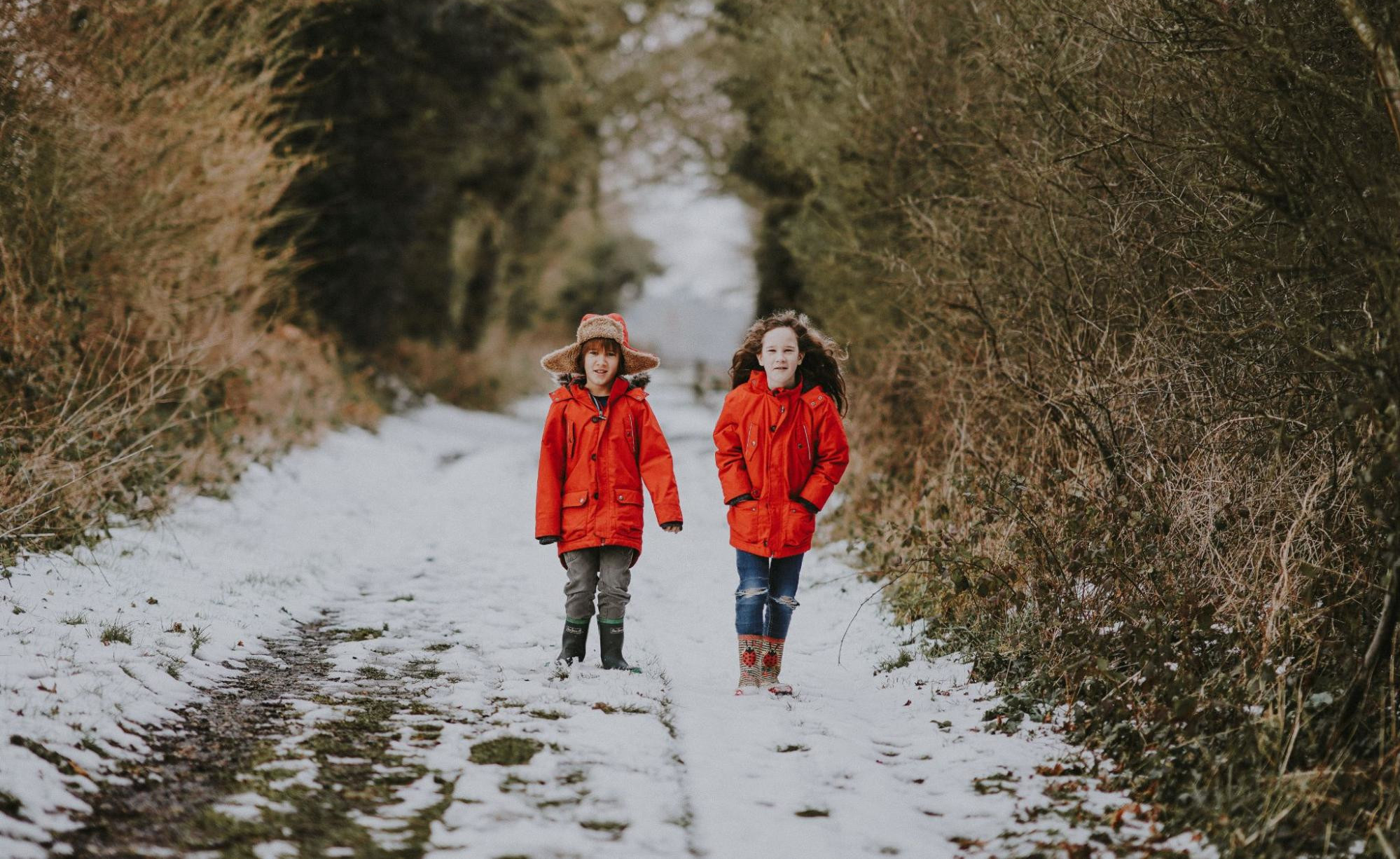 2 children dressed in red coats walk along a snowy trail